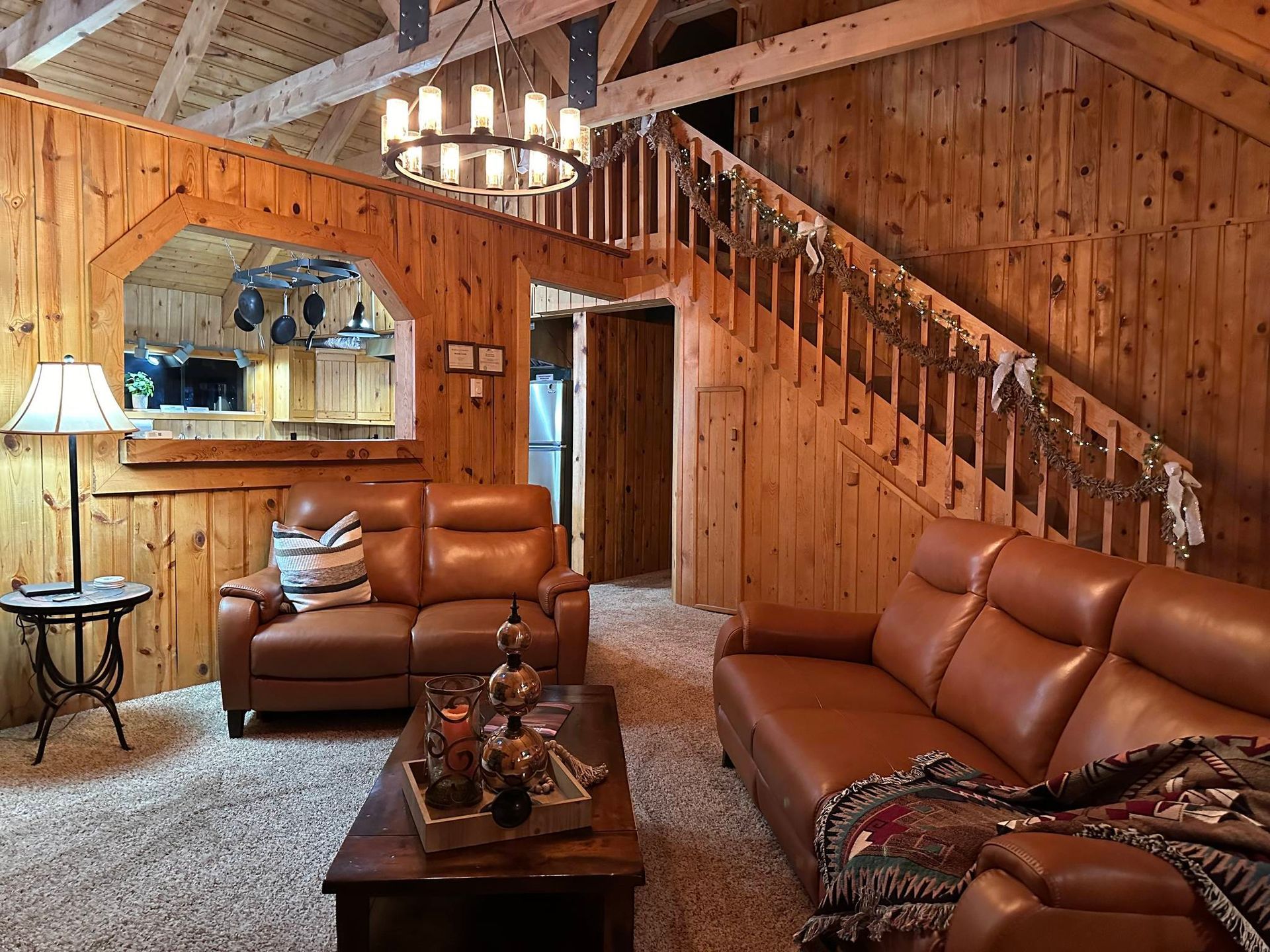 A rustic living room with wood paneled walls, two brown leather sofas, a coffee table, and a wooden staircase.