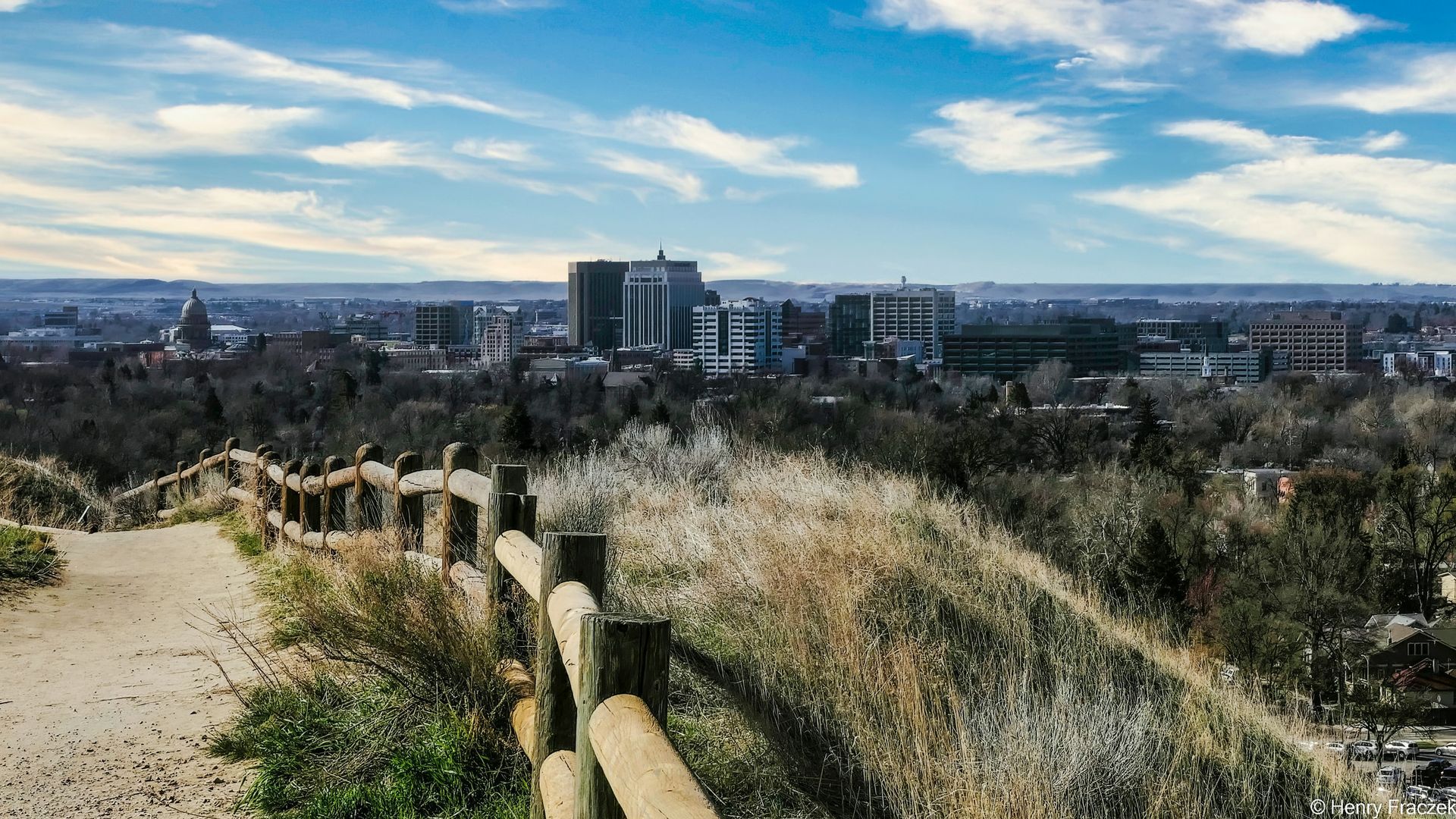 A wooden fence lines a trail on a grassy hillside, overlooking a city skyline under a blue sky with wispy clouds.