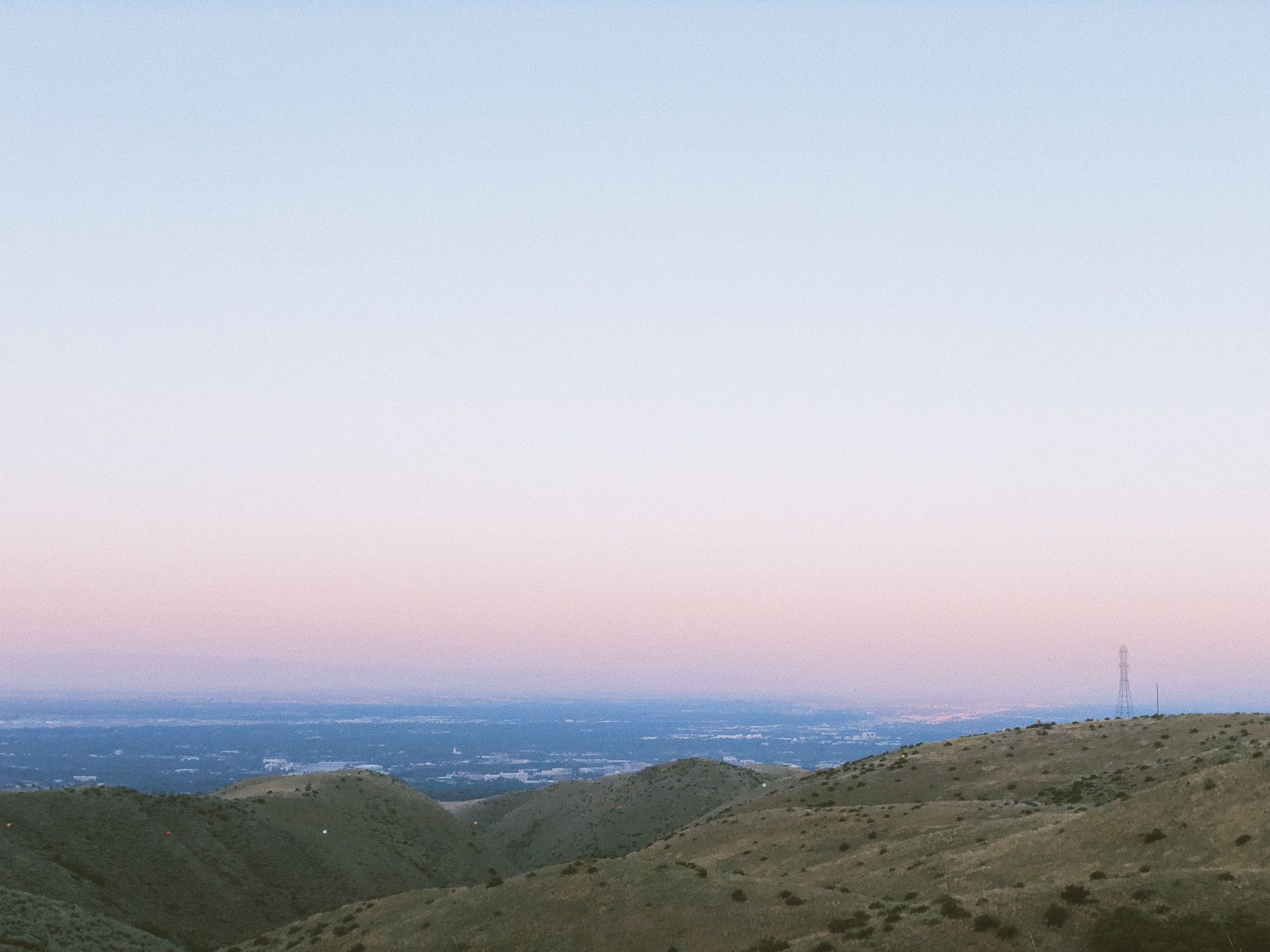A hazy, pink-tinted sunset sky illuminates a landscape of rolling, grassy hills overlooking a distant urban valley.