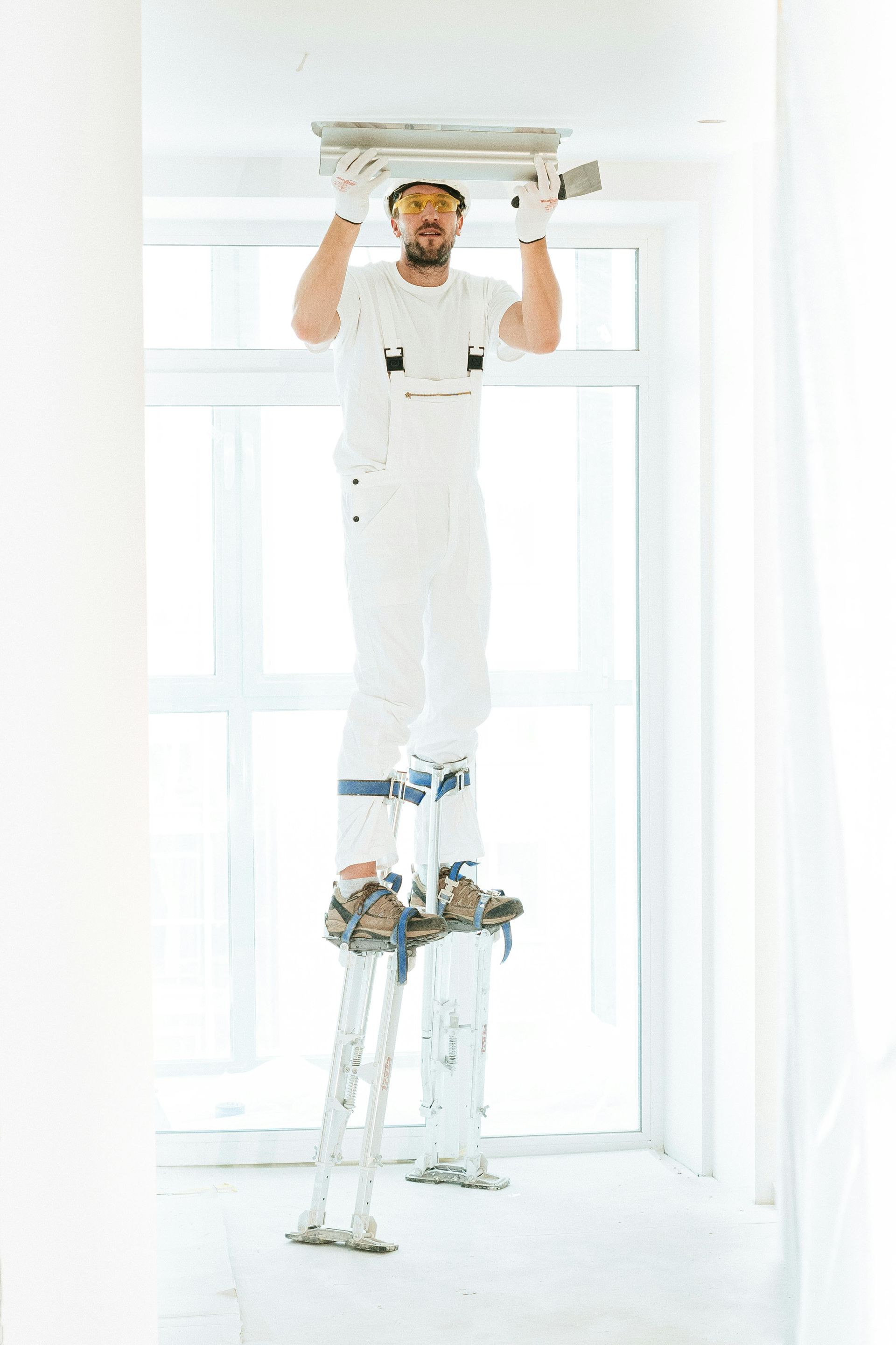 A construction worker in white clothing uses tall stilts to reach the ceiling to apply plaster with a trowel.