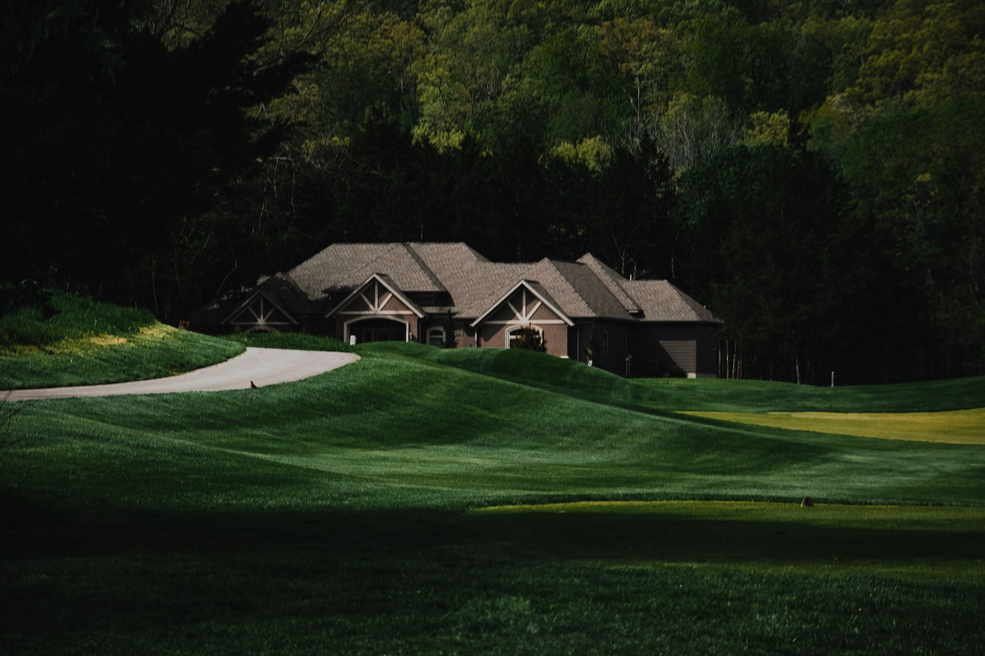 A large house sits at the edge of a lush, rolling green golf course surrounded by a dense forest under a moody sky.