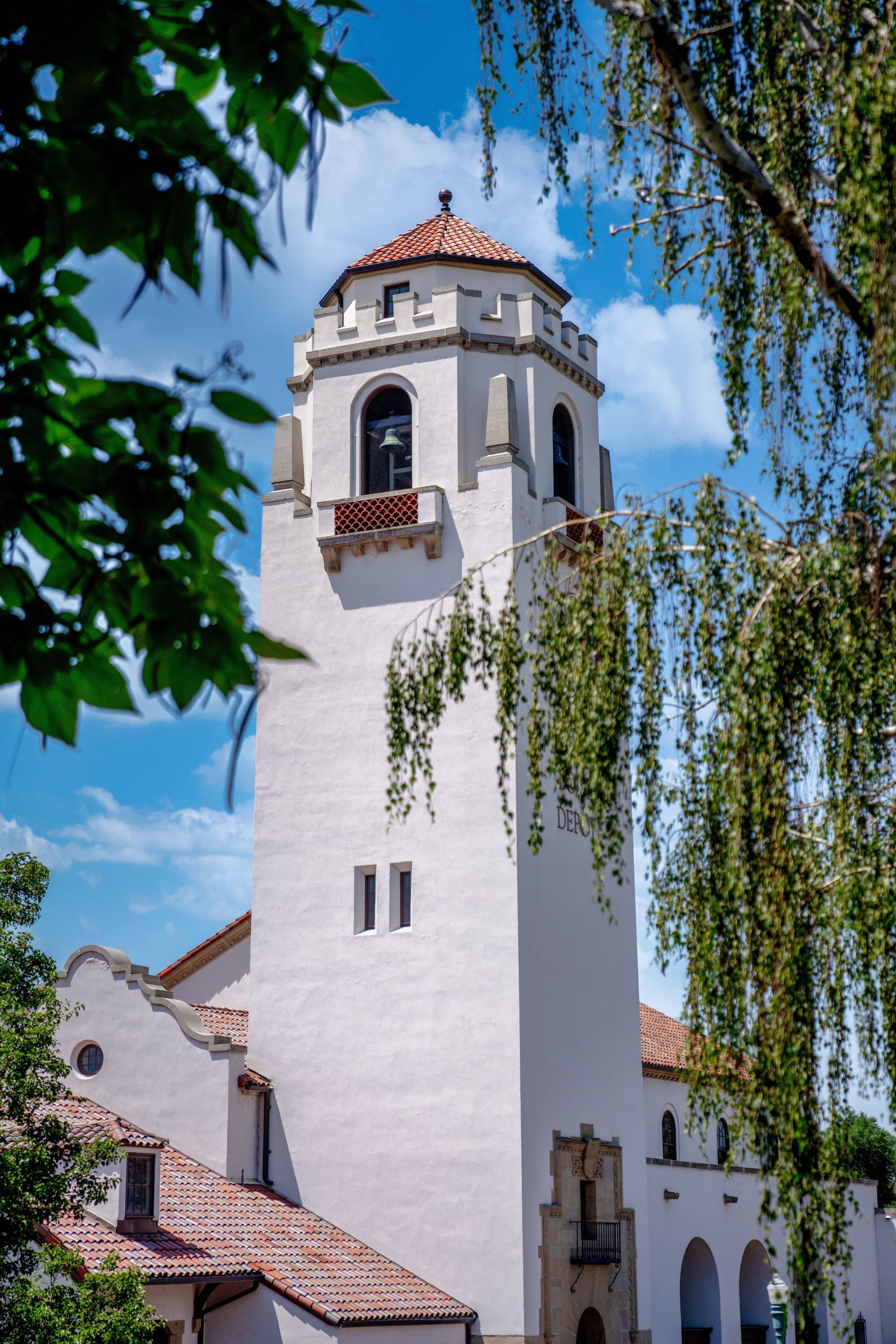 A white stucco tower with a red tile roof stands against a blue sky, framed by the green leaves of nearby trees.