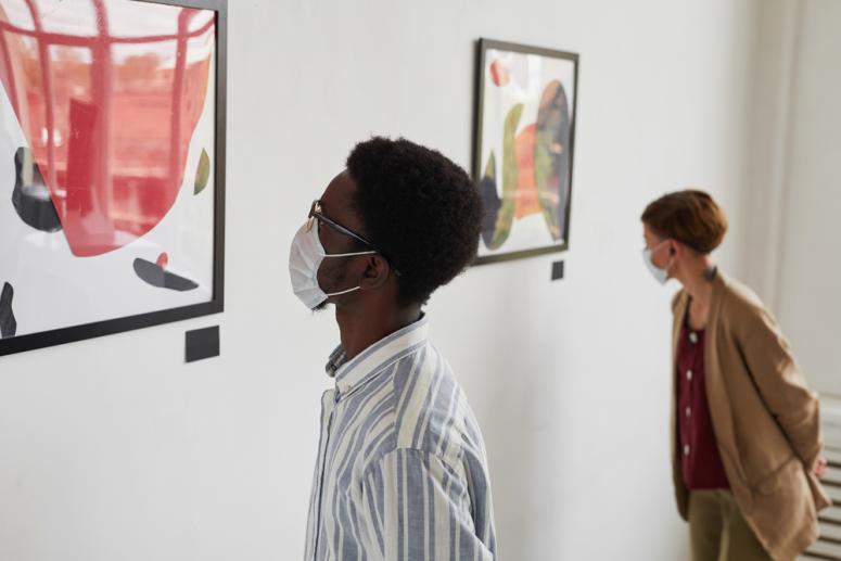 A man wearing a mask is looking at a painting in a museum.