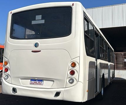 Vista traseira de um ônibus de passageiros branco com vidros fumê, estacionado ao ar livre em um dia ensolarado.