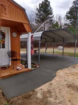 Brown shed with small porch and black chair, connected to a carport on a concrete pad.