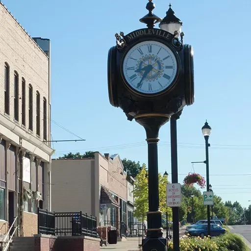 Black street clock on a sunny downtown street with buildings, lampposts, and parked cars in the background