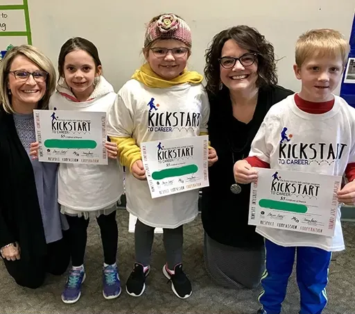 Five people posing indoors, three holding “Kickstart” fundraiser signs and smiling at the camera