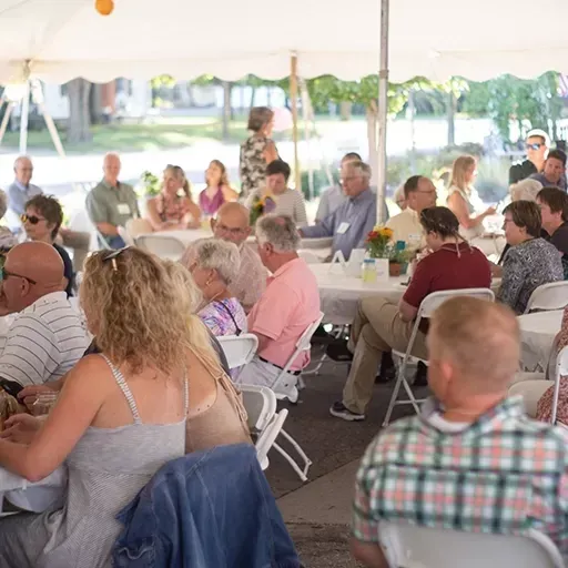 People seated at tables under a white tent at an outdoor gathering