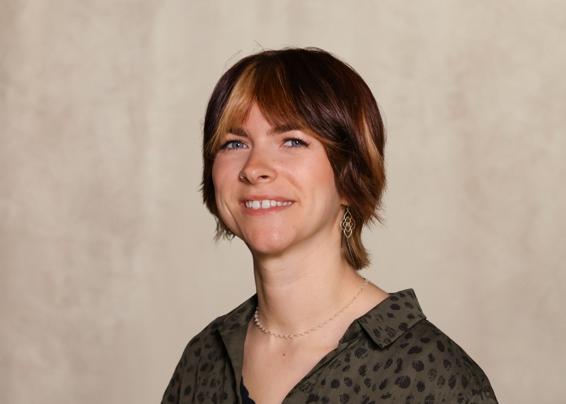 Portrait of a smiling person with short brown hair, wearing a dark patterned shirt against a beige backdrop
