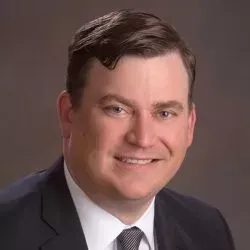 Studio headshot of a smiling man in a dark suit and tie against a gray background