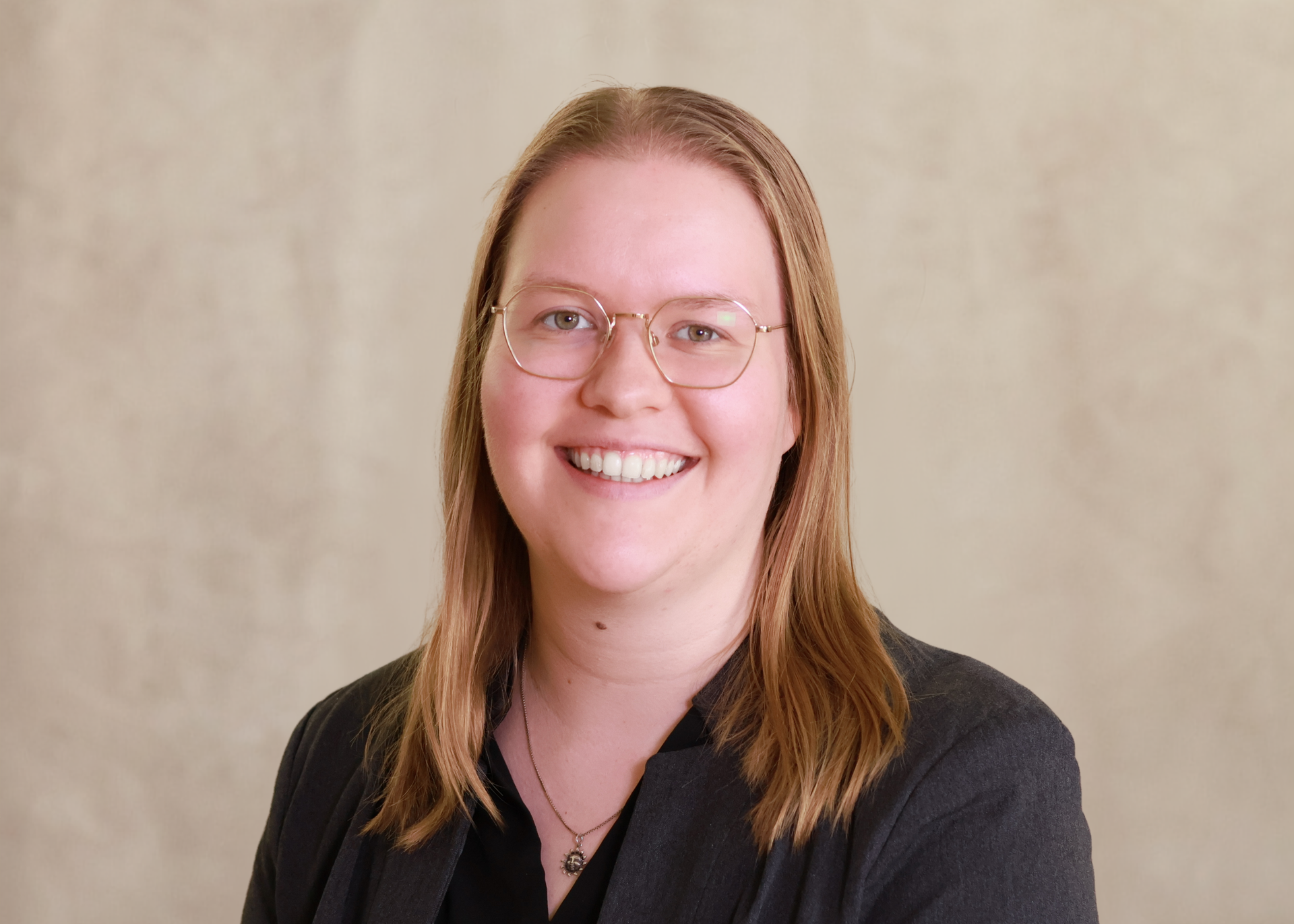 Smiling person with glasses and long hair, wearing a black blazer against a beige backdrop