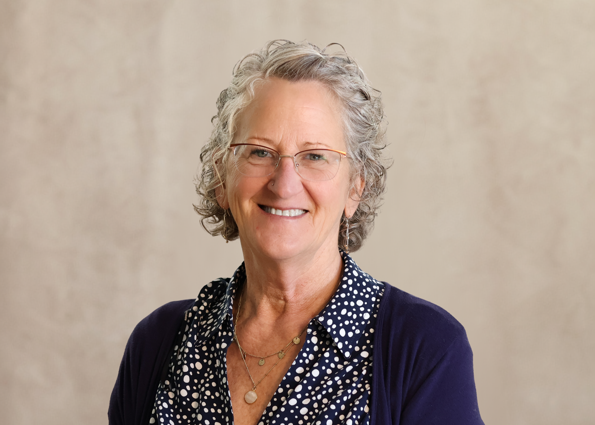 Smiling woman with curly gray hair and glasses, wearing a navy cardigan over a patterned blouse.