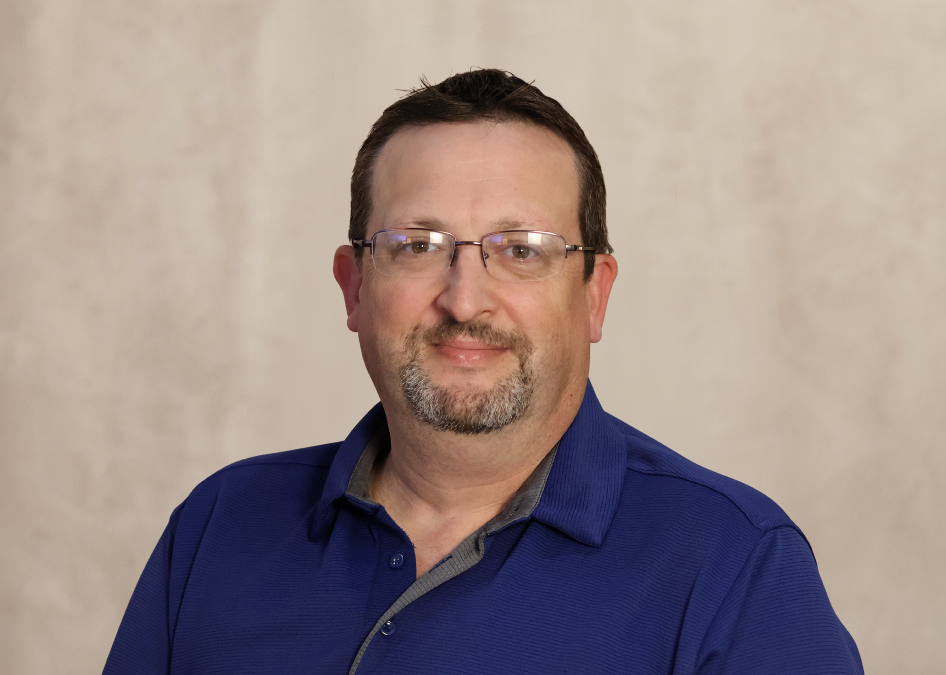 Man in glasses wearing a blue shirt against a beige backdrop, facing the camera with a slight smile
