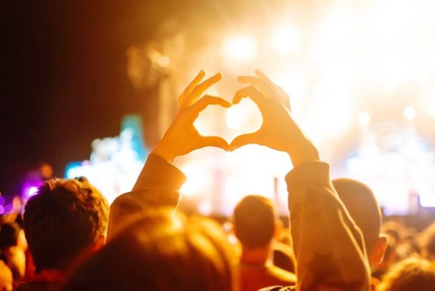 Crowd at a concert with hands forming a heart shape against bright stage lights
