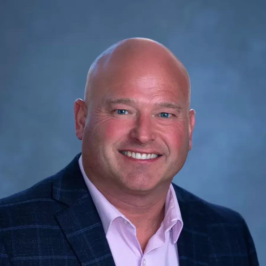 Smiling bald man in a dark blue plaid blazer against a soft blue studio background