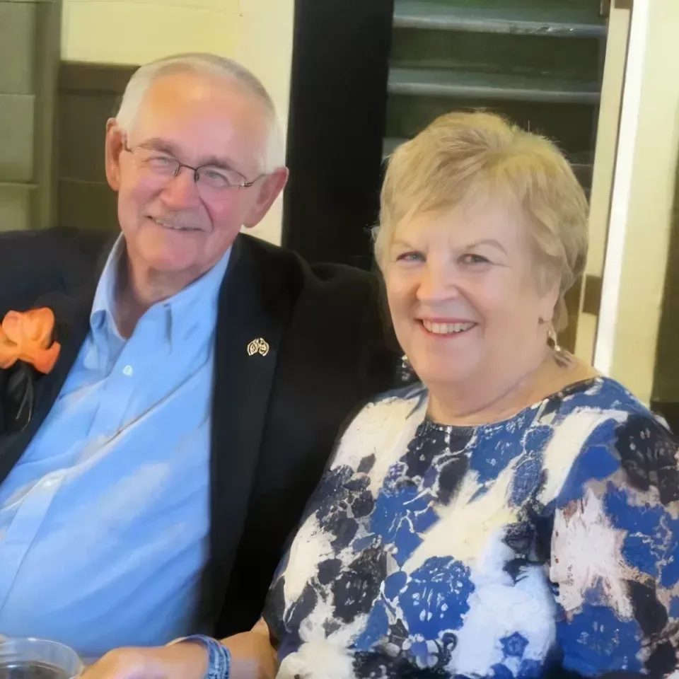Smiling couple seated indoors, the man in a dark jacket and blue shirt, the woman in a blue-and-white floral top.