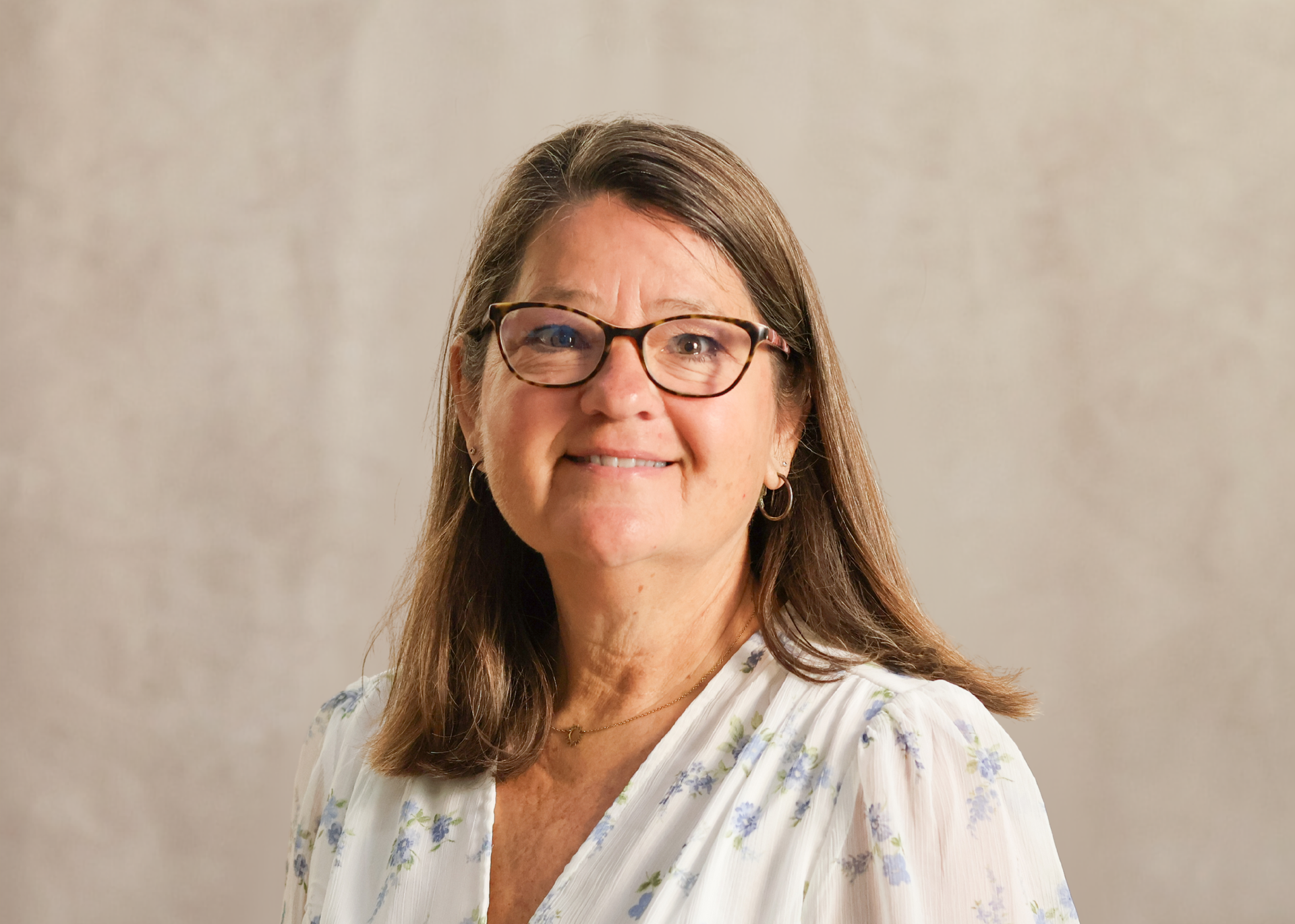 Smiling woman with glasses in a white floral blouse against a beige background