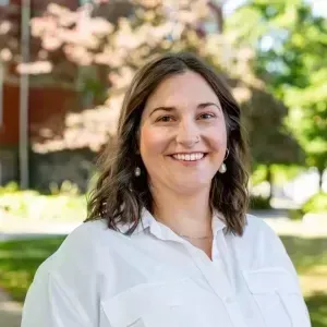 Smiling person in a white shirt standing outdoors with trees and a building in the background