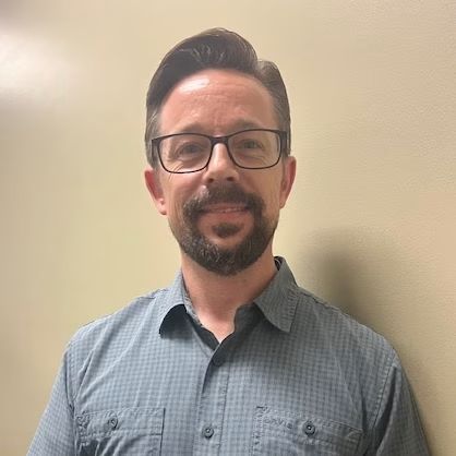 Man standing against a plain wall, wearing glasses and a blue button-up shirt, smiling slightly.