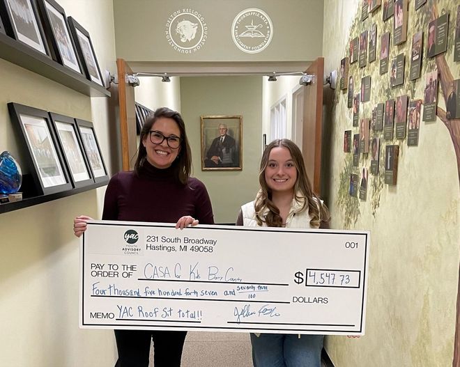 Two people smiling in a hallway hold a large ceremonial check.