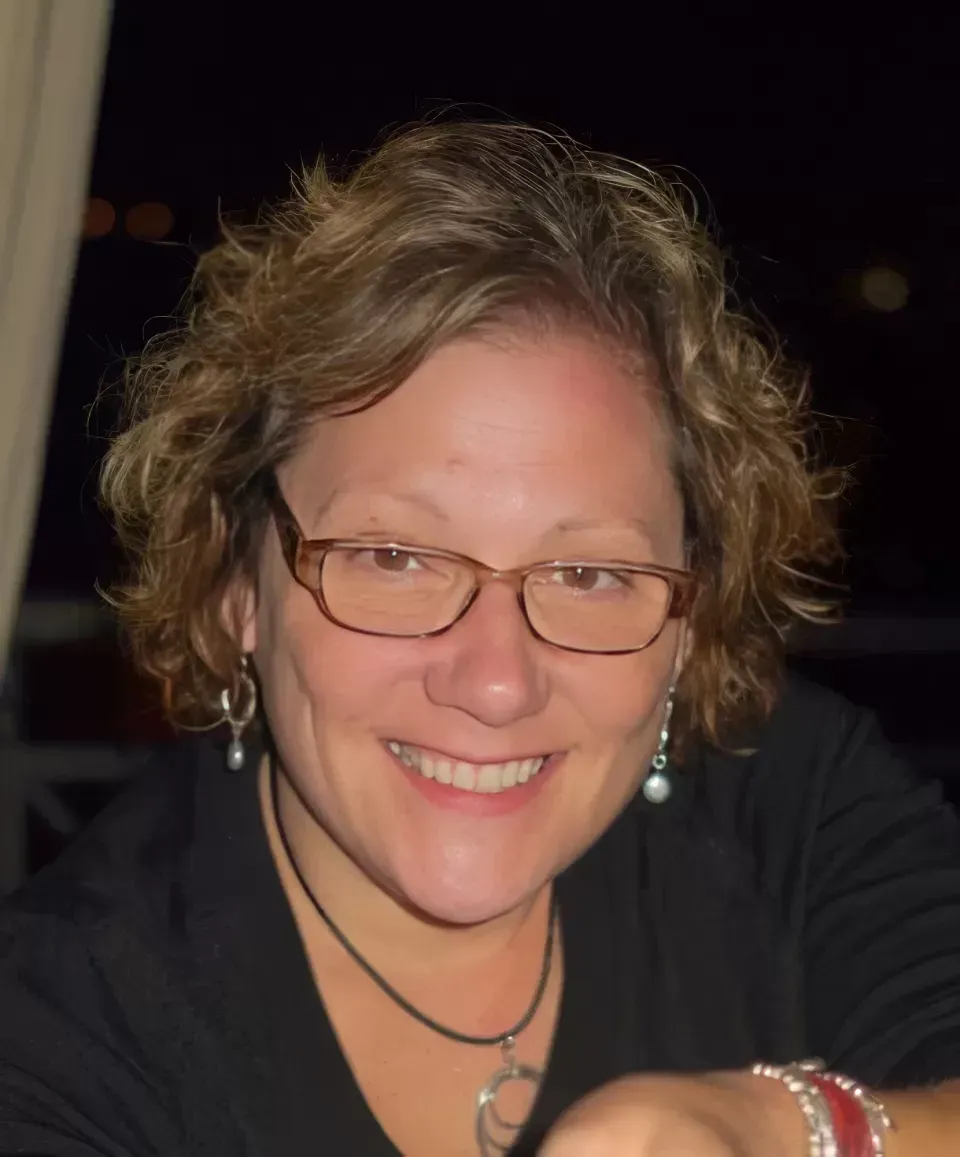 Smiling person with glasses and curly hair in a dark setting, wearing earrings and layered necklaces