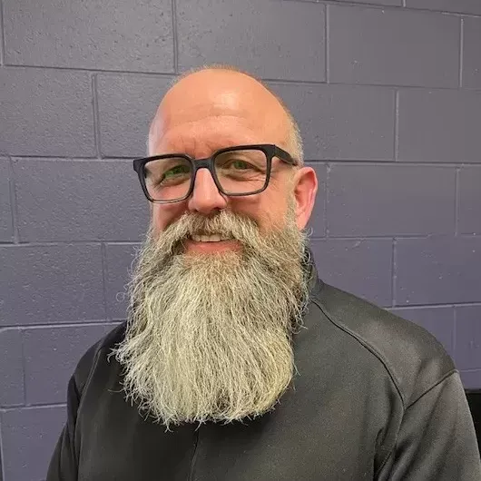 Man with glasses and a long gray beard standing against a gray brick wall