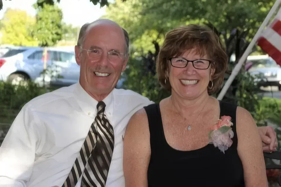 Two smiling adults seated outdoors at a gathering, with trees and parked cars in the background