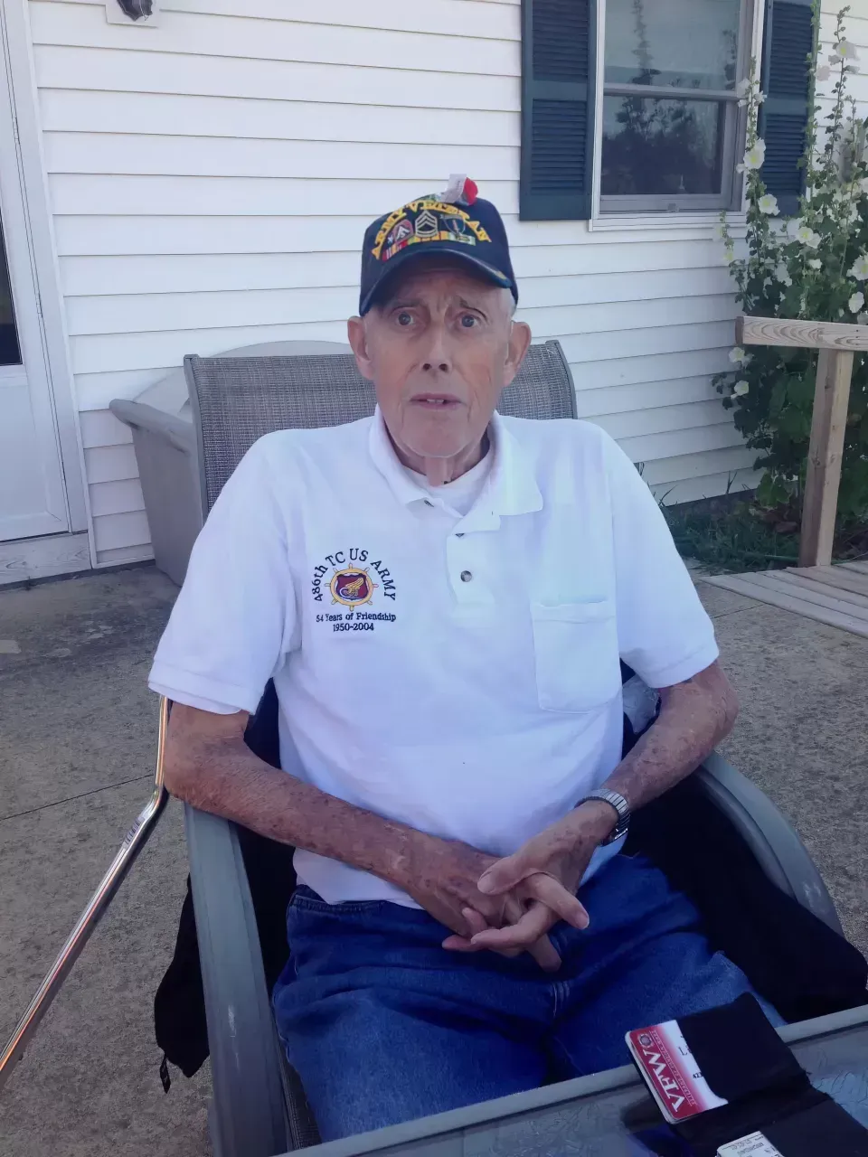 Man seated outdoors in a wheelchair on a porch, wearing a white polo shirt and colorful cap.