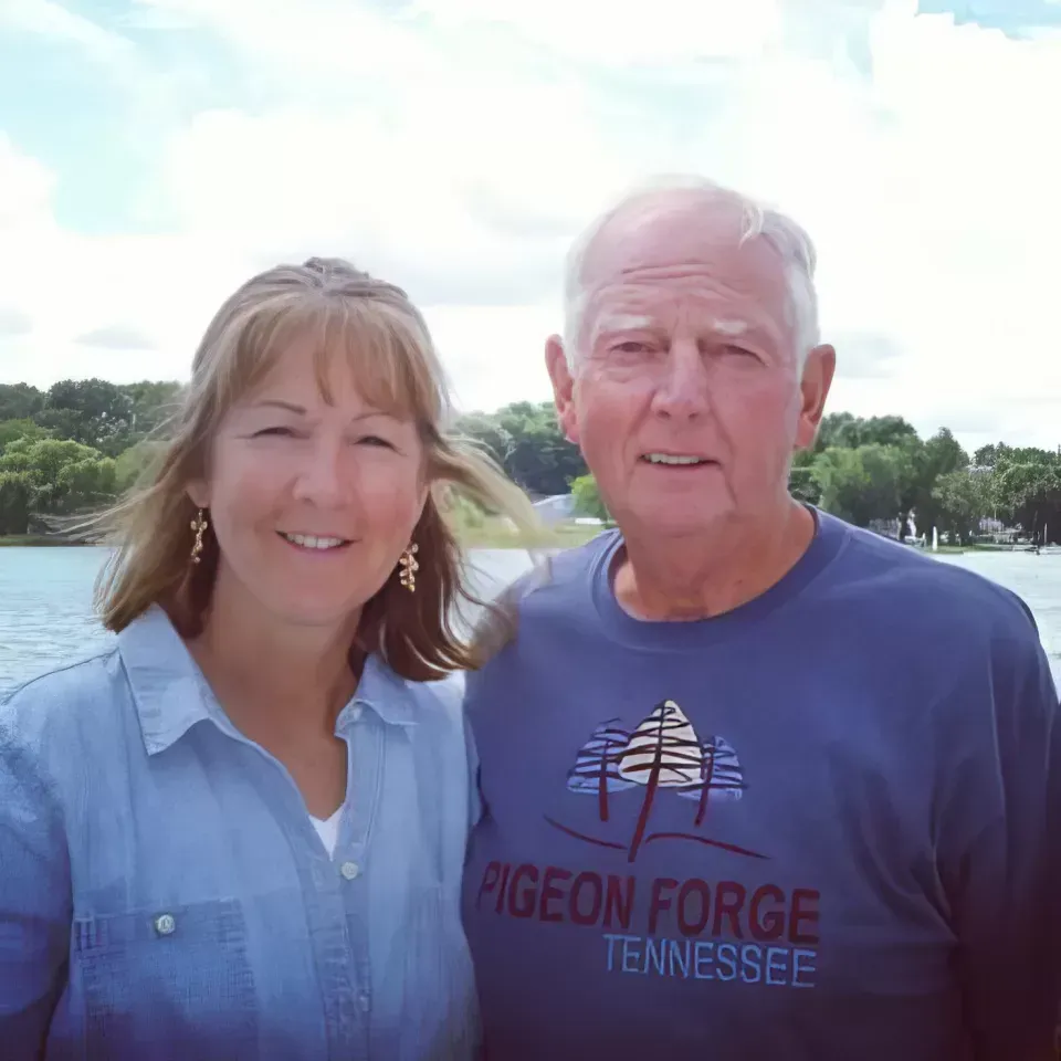 Two people smiling outdoors by a lake, with trees and a blue sky in the background.
