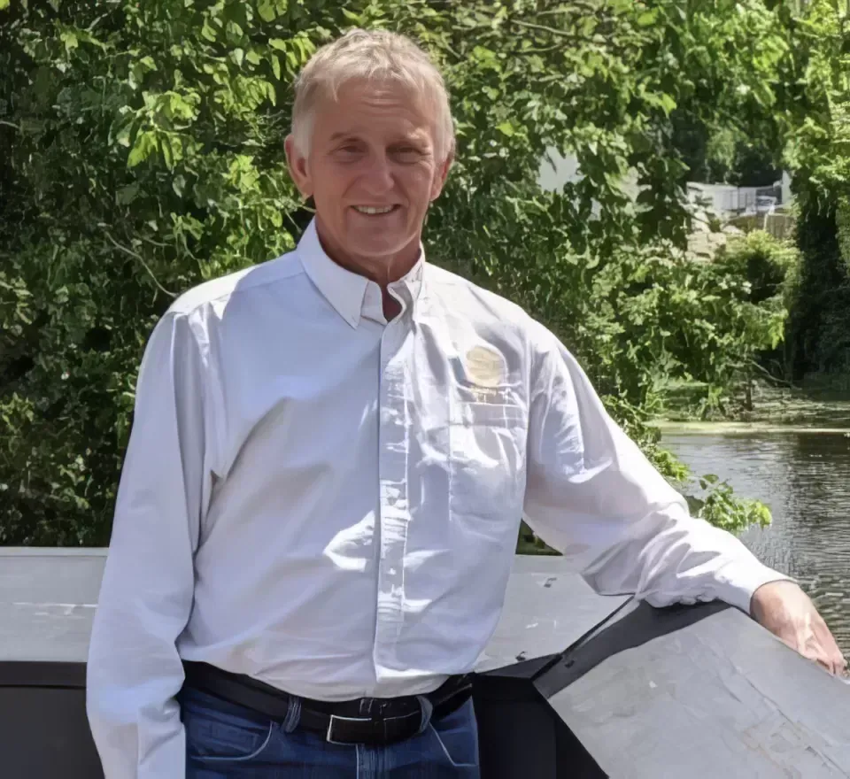 Man standing outdoors by a railing in a white shirt, with green trees in the background