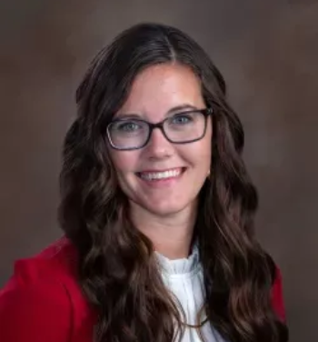 Portrait of a smiling person with glasses and wavy hair, wearing a red blazer against a brown backdrop