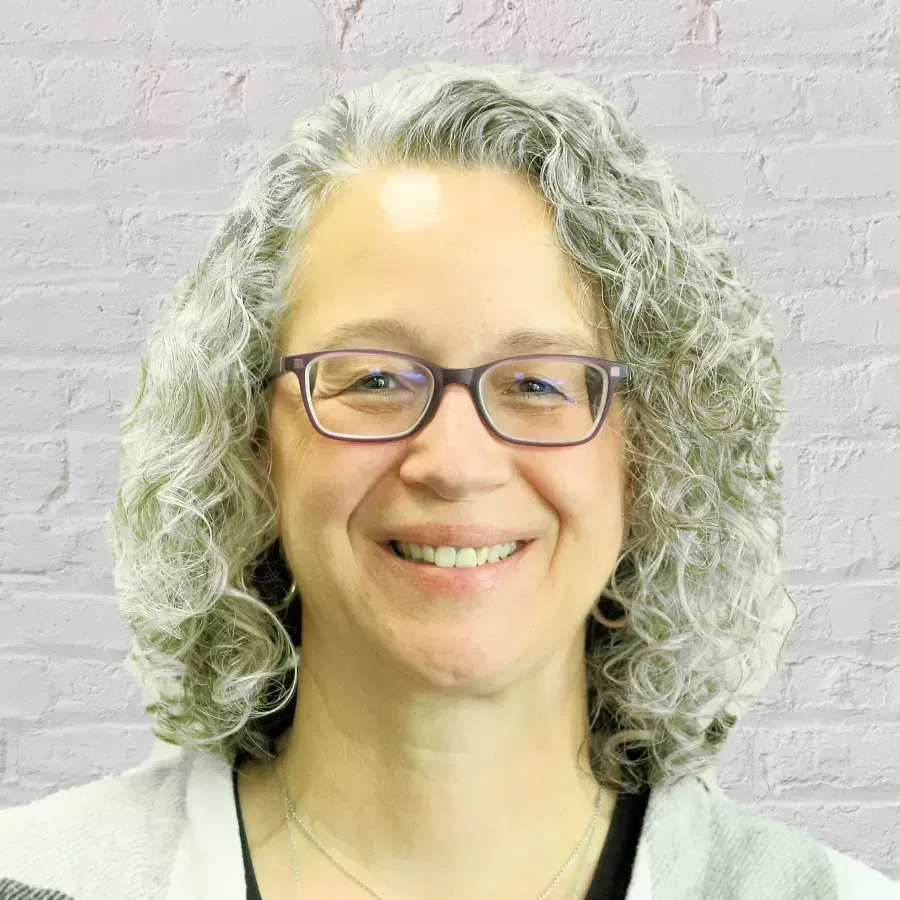 Smiling person with curly gray hair and glasses against a light brick wall
