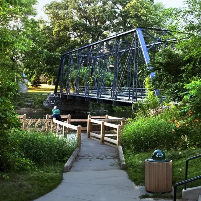 Blue steel bridge over a stream in a leafy park, with a concrete path and wooden railings in the foreground