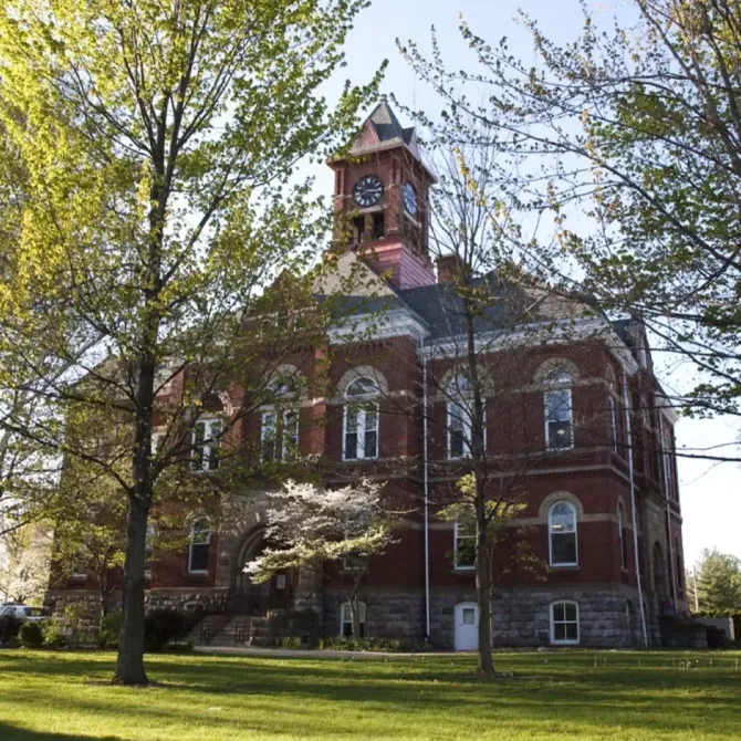 Red brick historic building with a small tower, seen through leafy trees on a grassy lawn