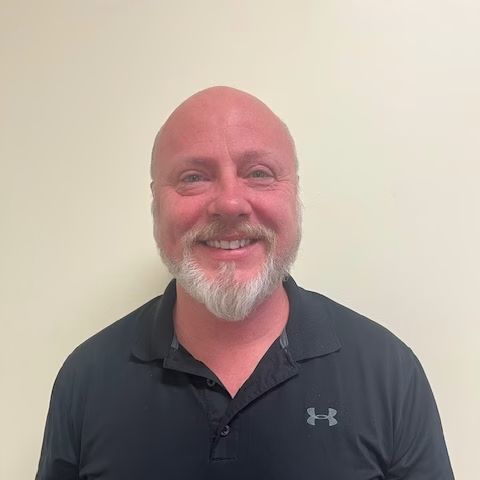 Smiling bald man with a white beard wearing a dark polo shirt against a plain wall