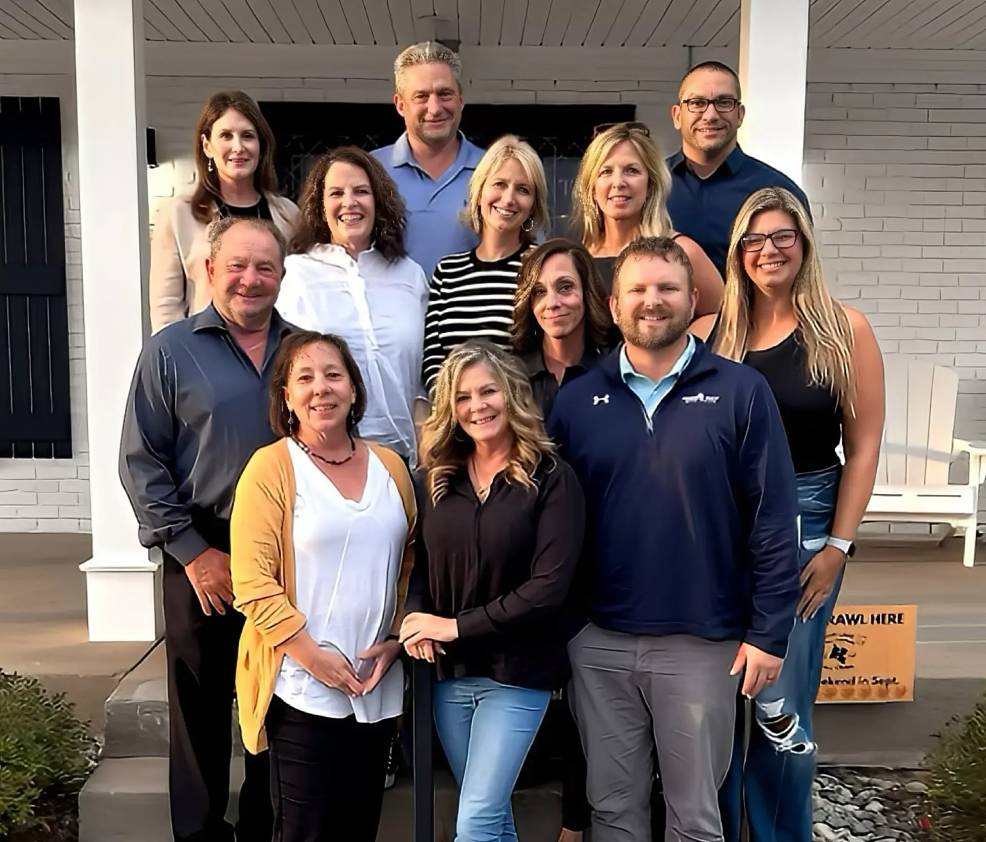 Group of eleven people posing on a front porch, smiling in casual clothes.