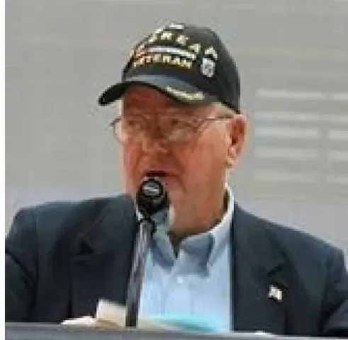 Man speaking at a microphone, wearing a dark blazer and black cap with text.