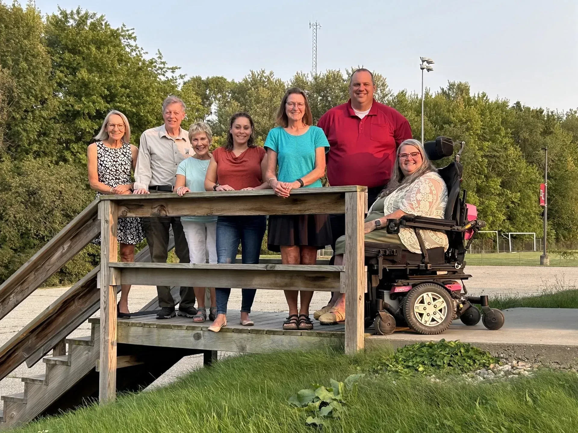 Group posing on a wooden bridge in a park beside a person on a mobility scooter