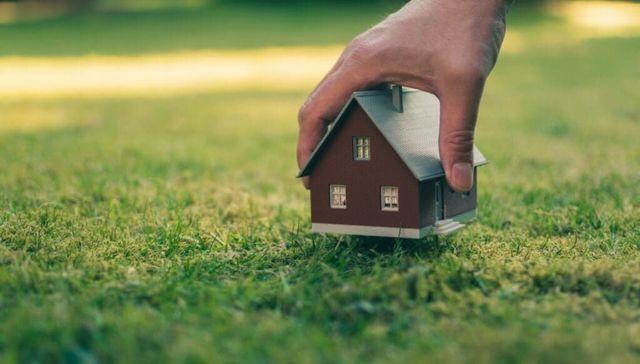 Hand placing a small house model on green grass, symbolizing home or property