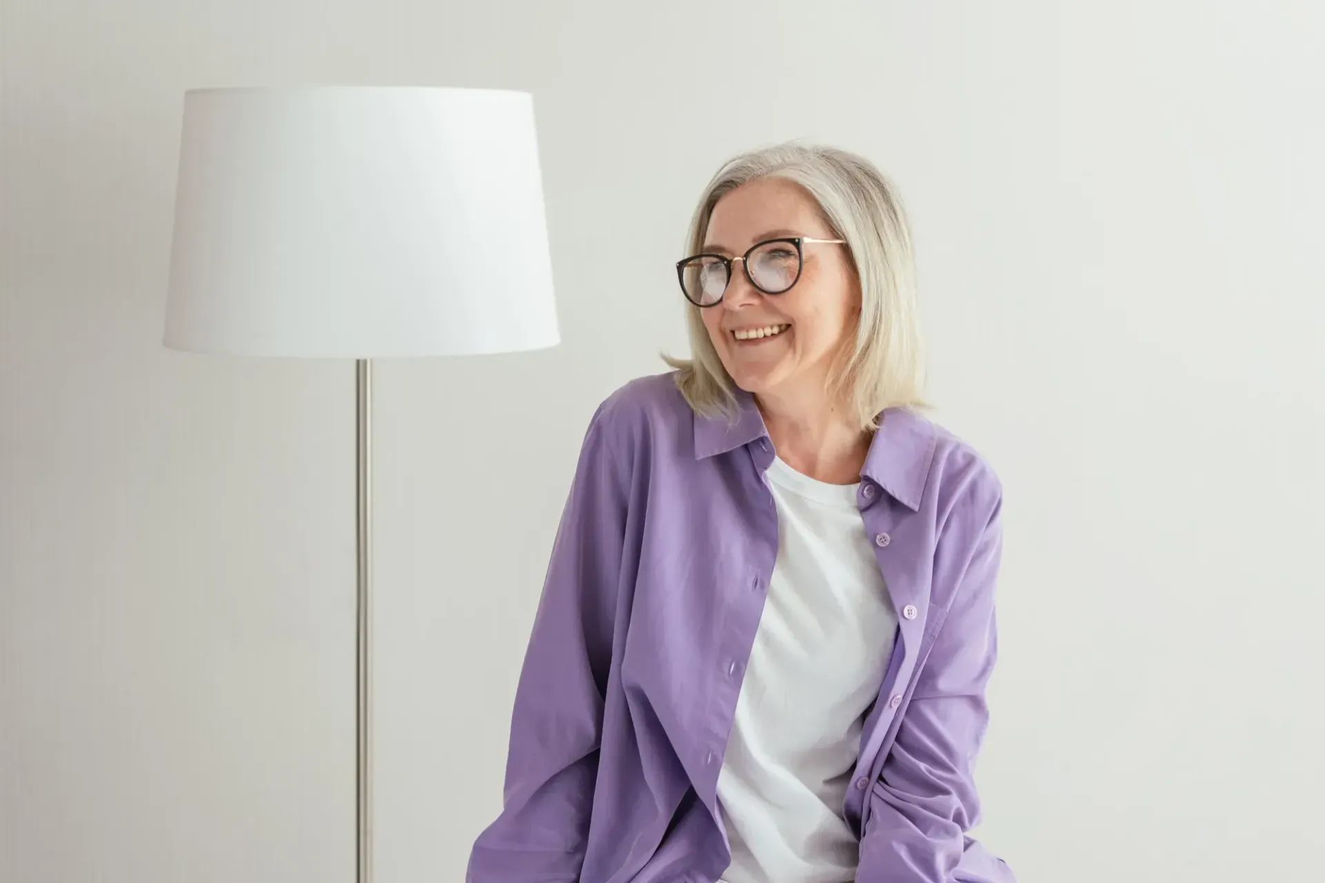Smiling person in a purple shirt beside a white floor lamp against a light wall