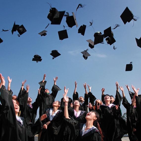 Graduates in black gowns toss mortarboards into the blue sky at an outdoor ceremony