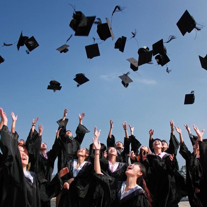 Graduates in black gowns toss caps into a blue sky