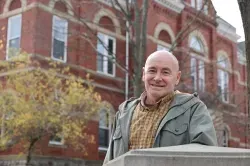 Smiling man in a green jacket standing by a stone railing in front of a red brick building