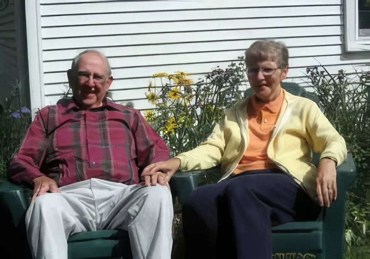 Two people sitting outdoors on green chairs, holding hands, with a house wall and flowers behind them