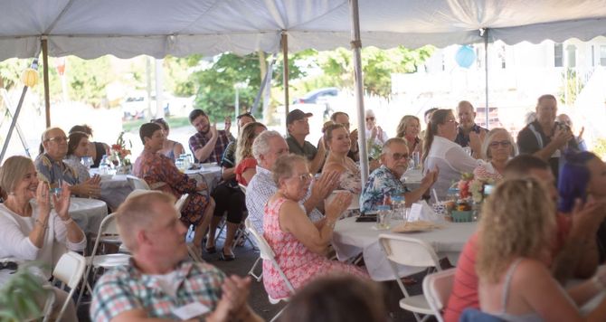 People seated at tables under a white tent, enjoying a group gathering or meal.