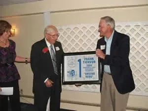 Two men in suits hold a framed award while a woman stands beside them at an indoor event