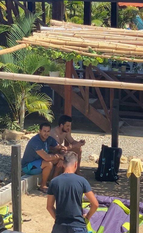 A group of men are sitting under a wooden structure on a beach.