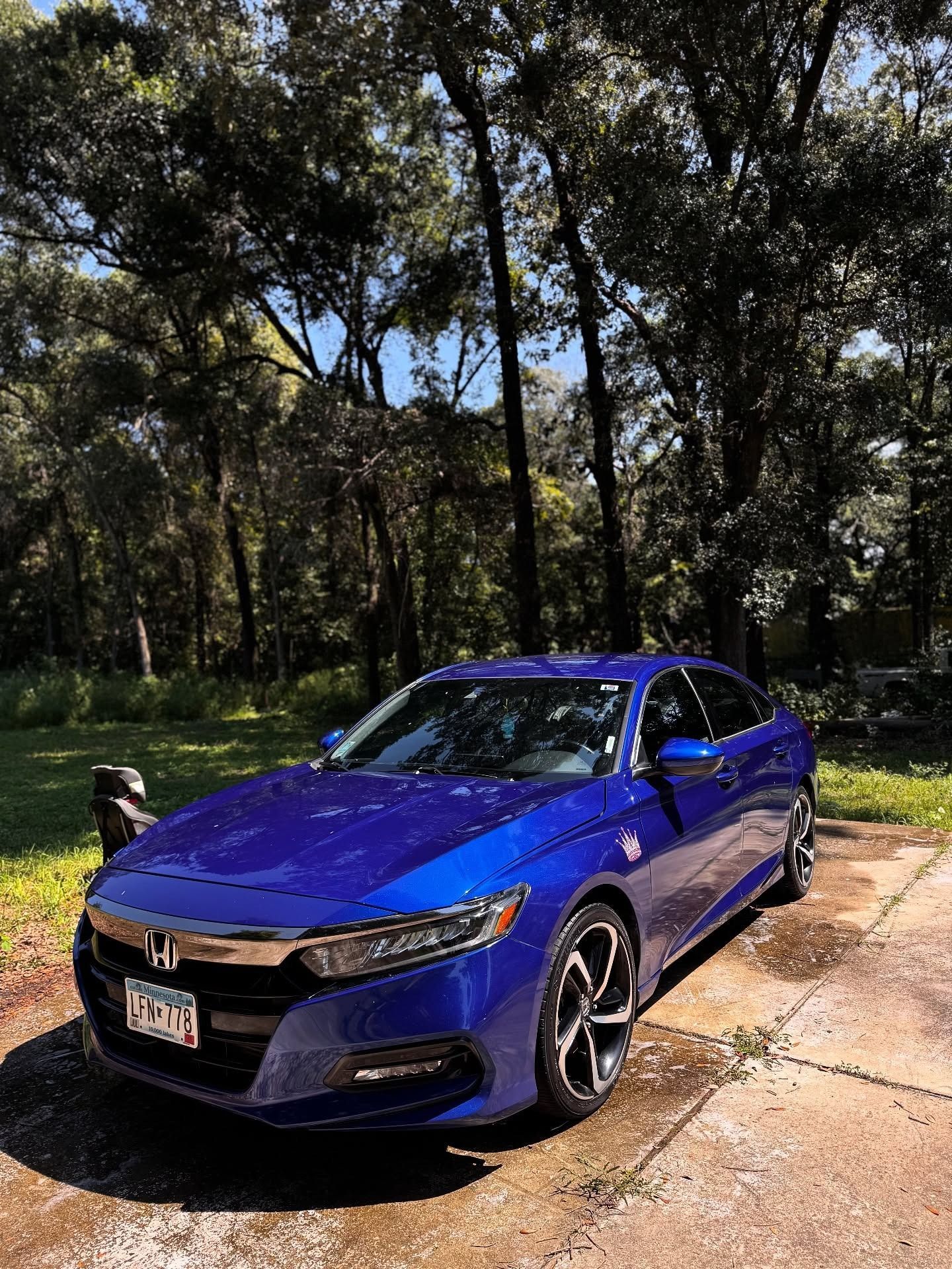 Blue sedan parked on a driveway beside trees in bright sunlight