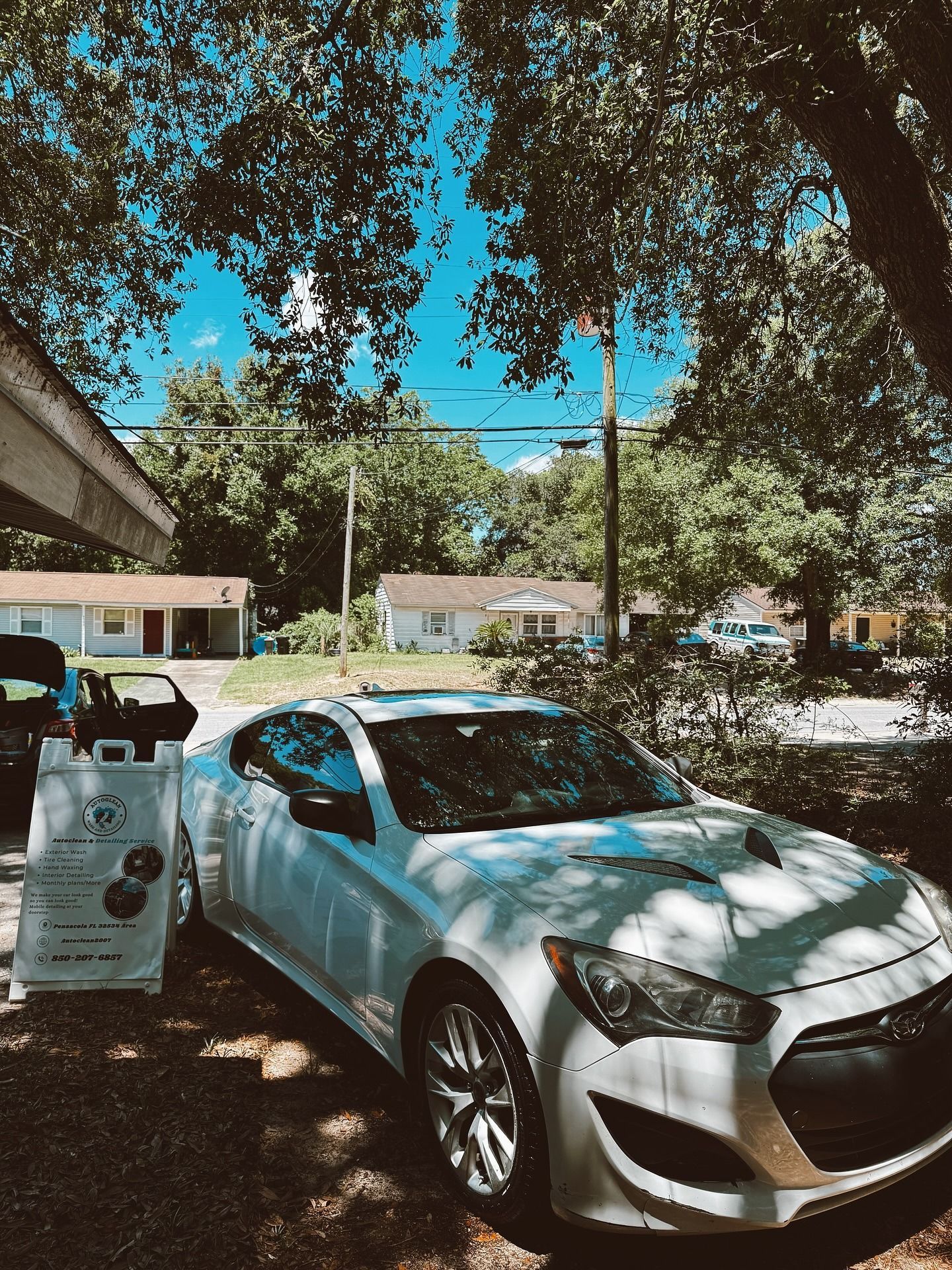 Silver car parked under trees in a shady lot beside a sidewalk sign, with houses in the background
