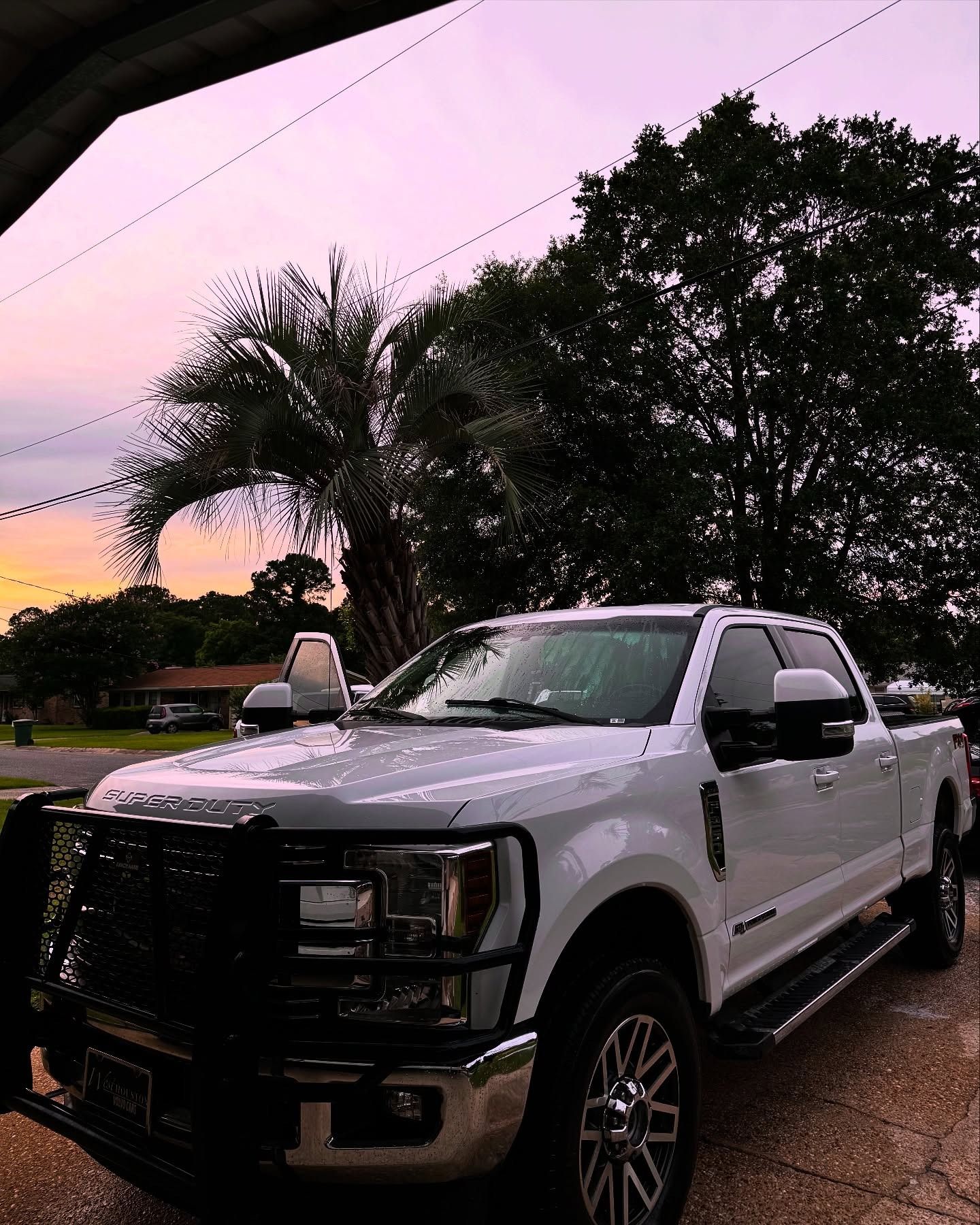 White pickup truck parked on a wet driveway at dusk under pink-purple sky, with trees in the background